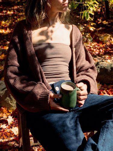Woman in a brown cardigan holding a green mug of cocoa among fallen autumn leaves